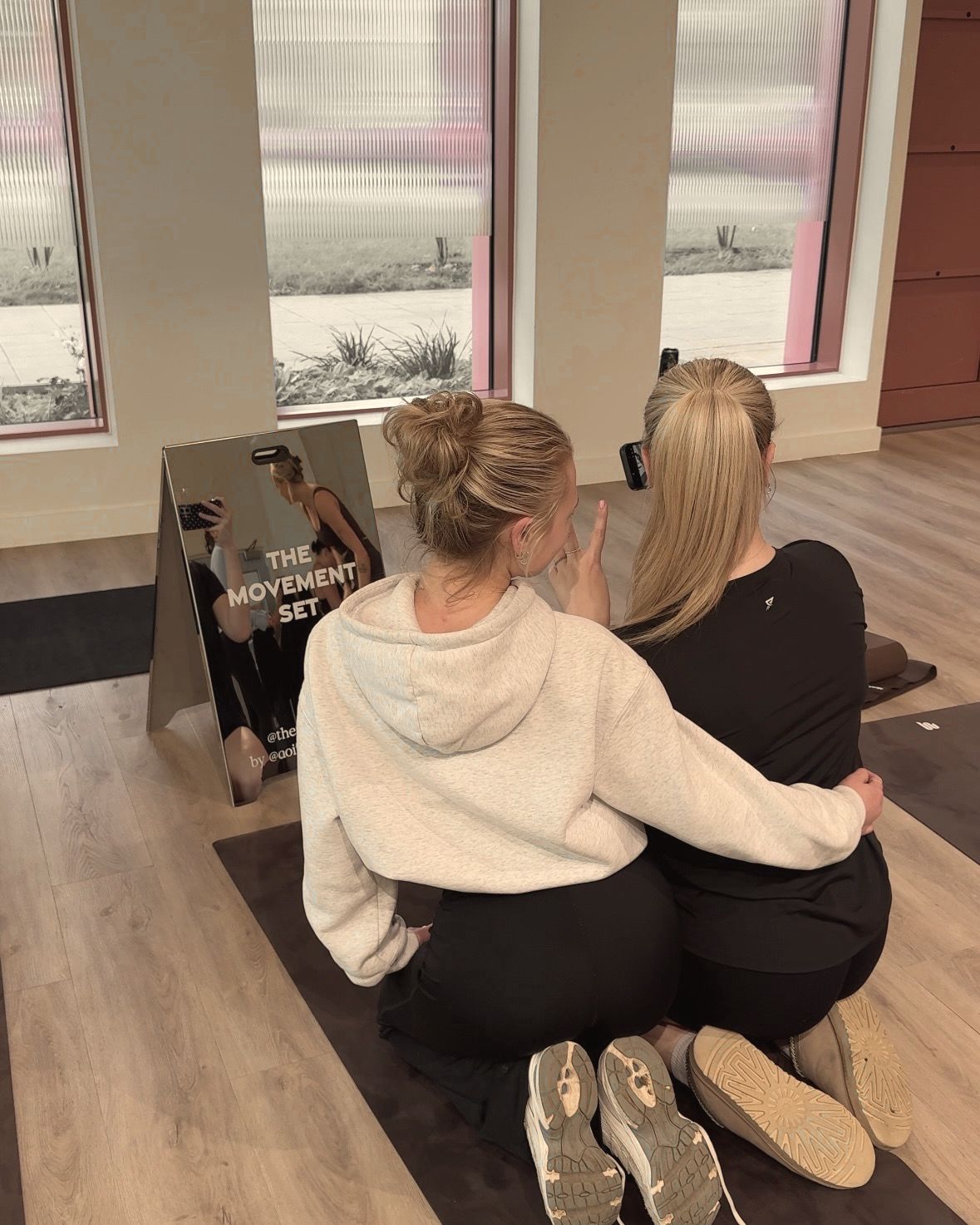 Two friends sitting together on a mat, taking a photo in The Movement Set mirror